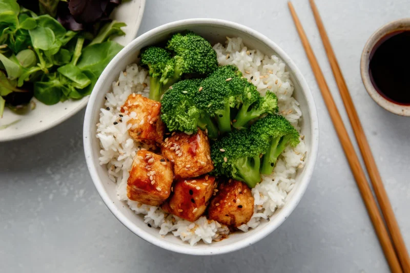Bowl of rice with broccoli and glazed tofu topped with sesame seeds, accompanied by chopsticks and soy sauce on a gray surface