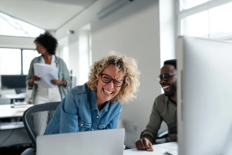 Woman laughing with colleague at laptop in bright office workspace