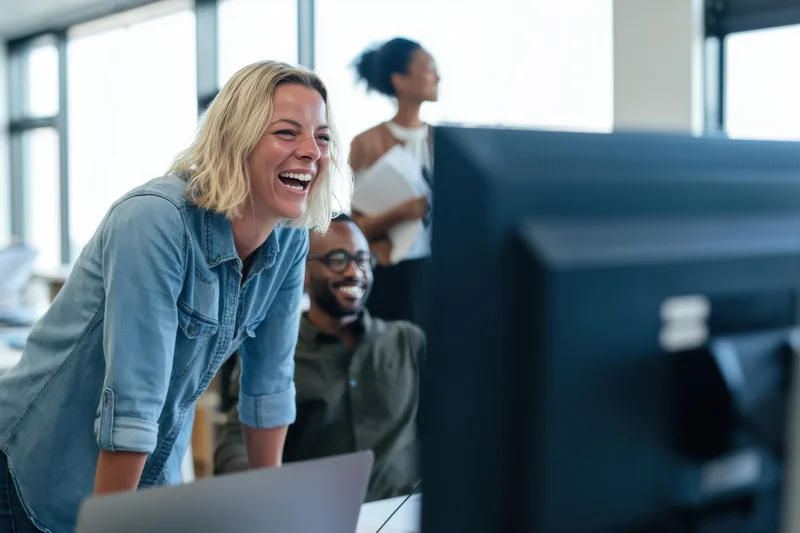 Blonde woman laughing while leaning over a laptop in a bright office with colleagues in the background