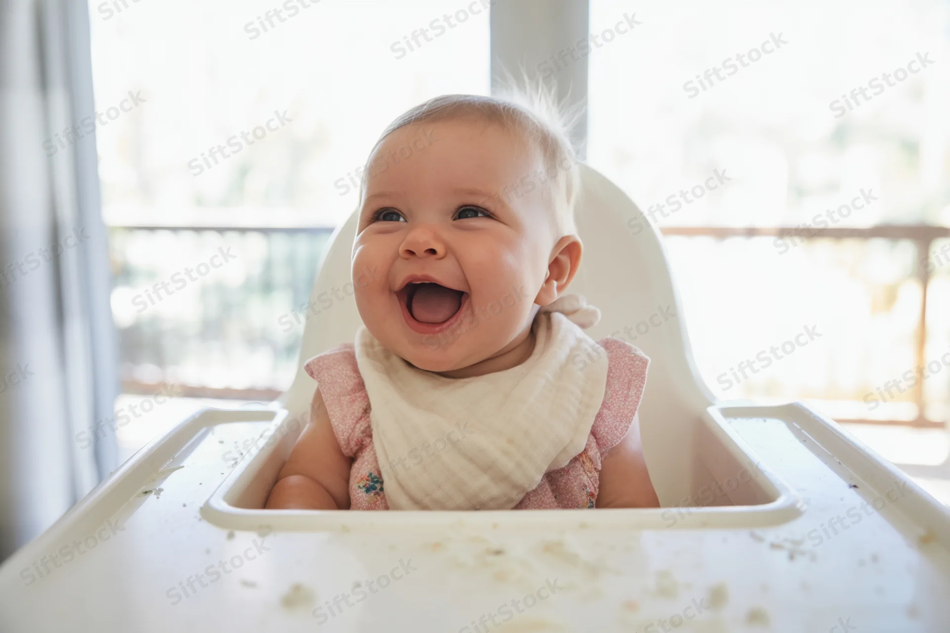 Baby smiling in high chair with crumbs on tray near window