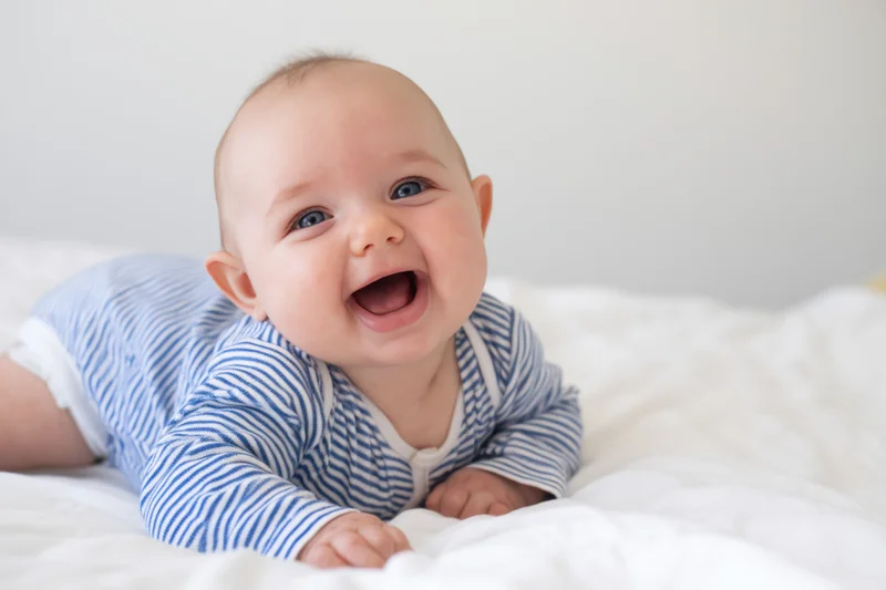 Baby crawling on white bedding wearing blue striped onesie, smiling broadly