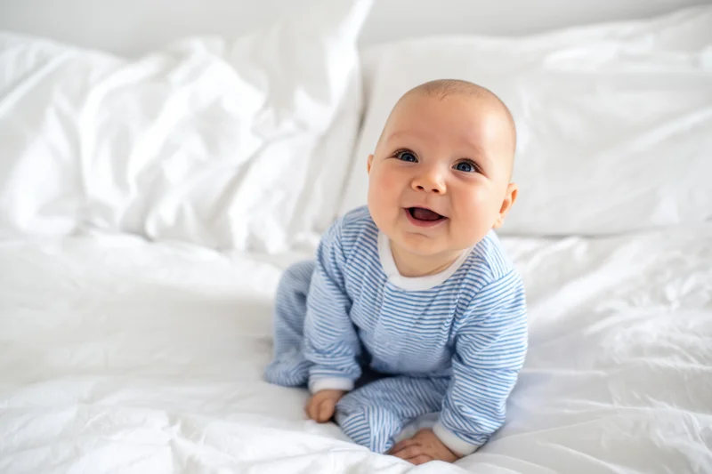 Baby sitting on white bed smiling in blue striped onesie