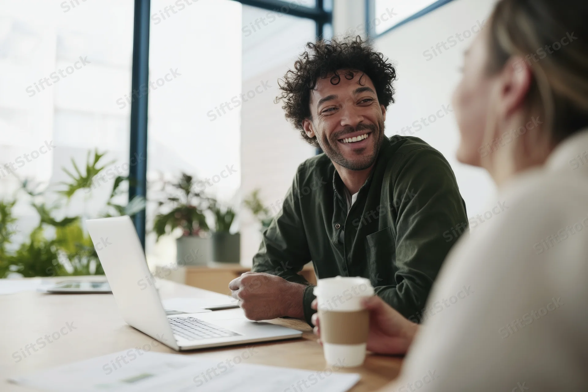 Smiling man in dark green shirt chatting with woman at office desk with laptop and coffee
