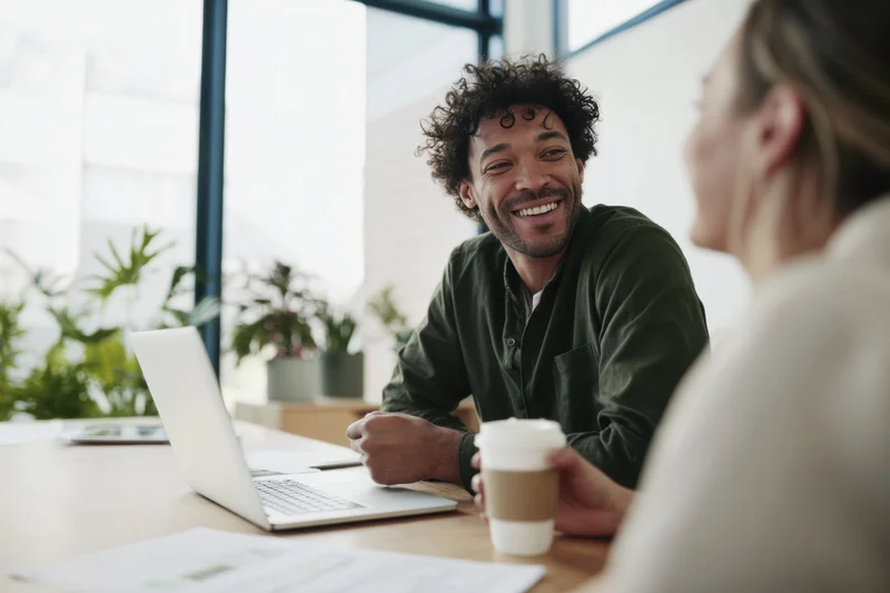 Smiling man in dark green shirt chatting with woman at office desk with laptop and coffee