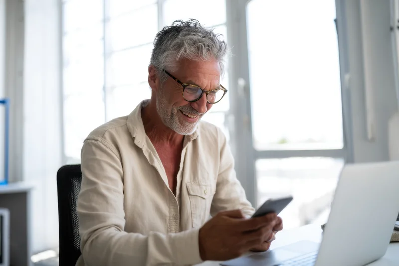 Middle-aged man smiling while using smartphone at desk with laptop in bright office