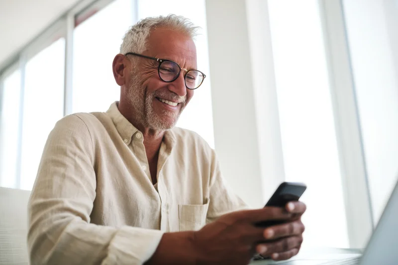 Smiling senior man using smartphone indoors near window