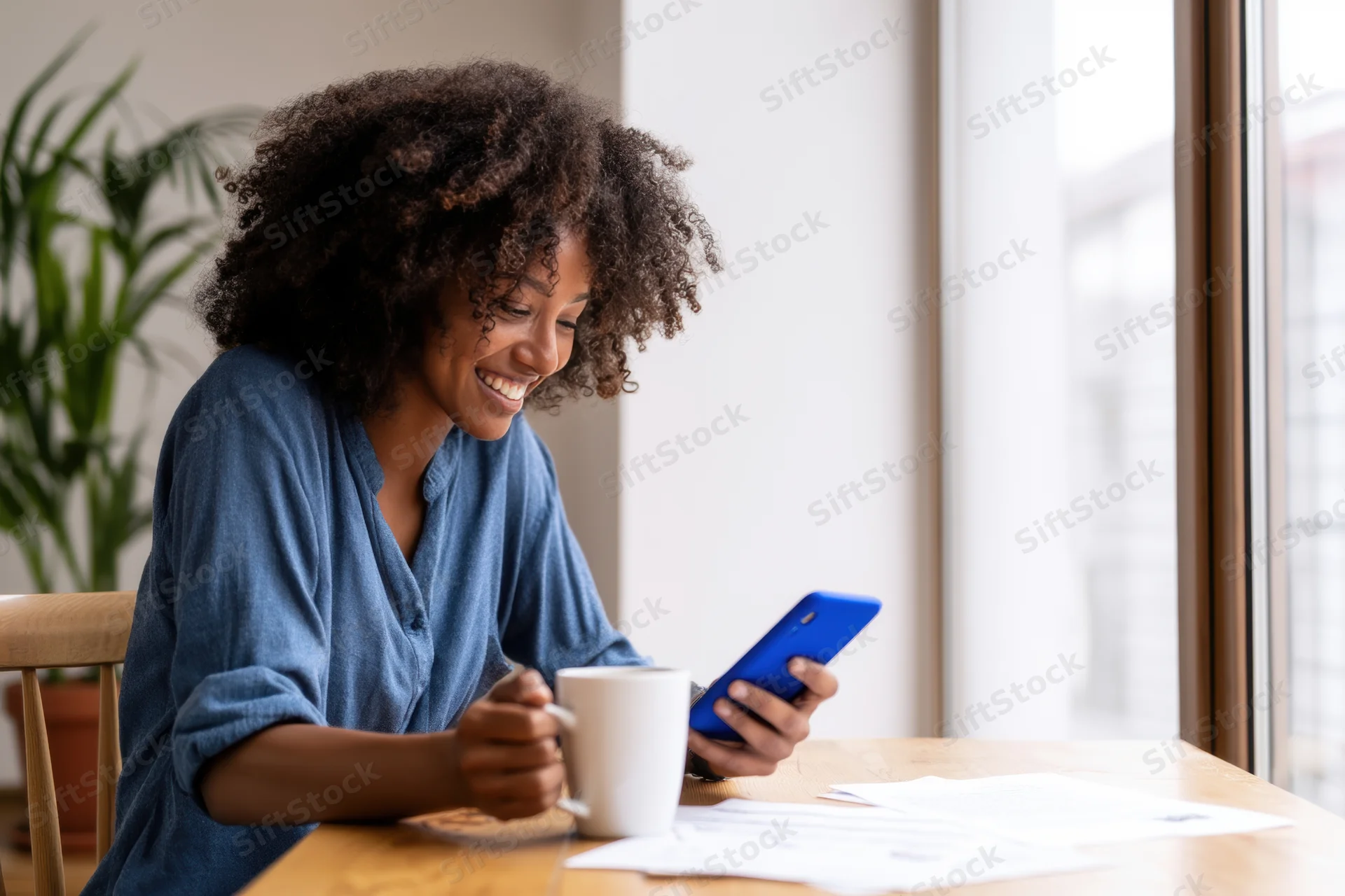 Woman smiling while using smartphone at wooden desk by window with coffee mug and papers