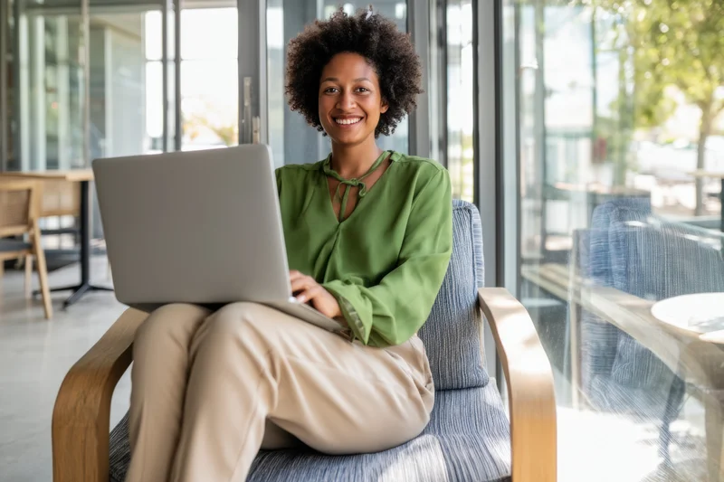 Woman smiling while using laptop on chair in modern cafe interior