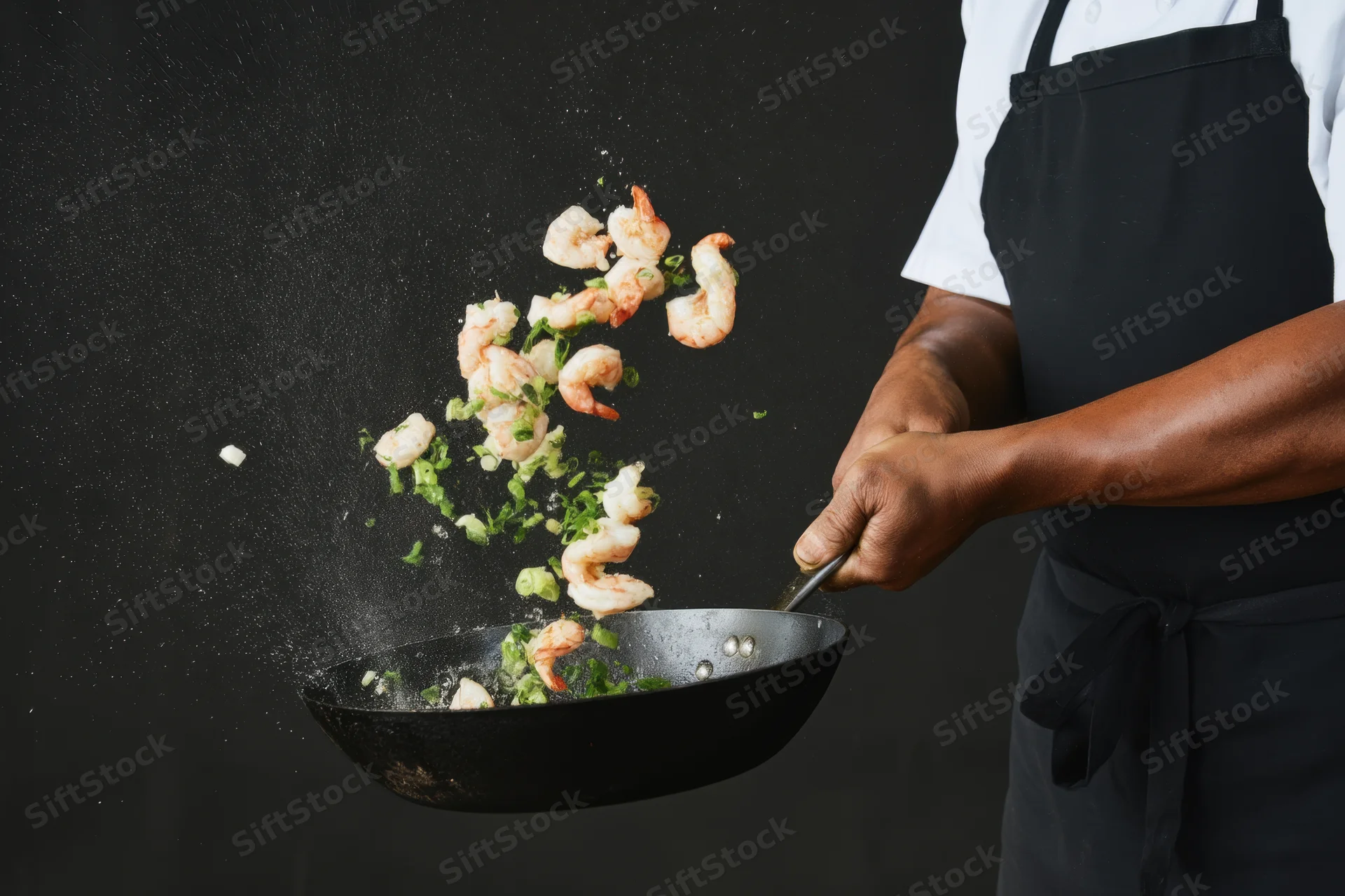 Chef tossing shrimp and green onions in a wok against black background