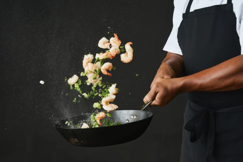 Chef tossing shrimp and green onions in a wok against black background