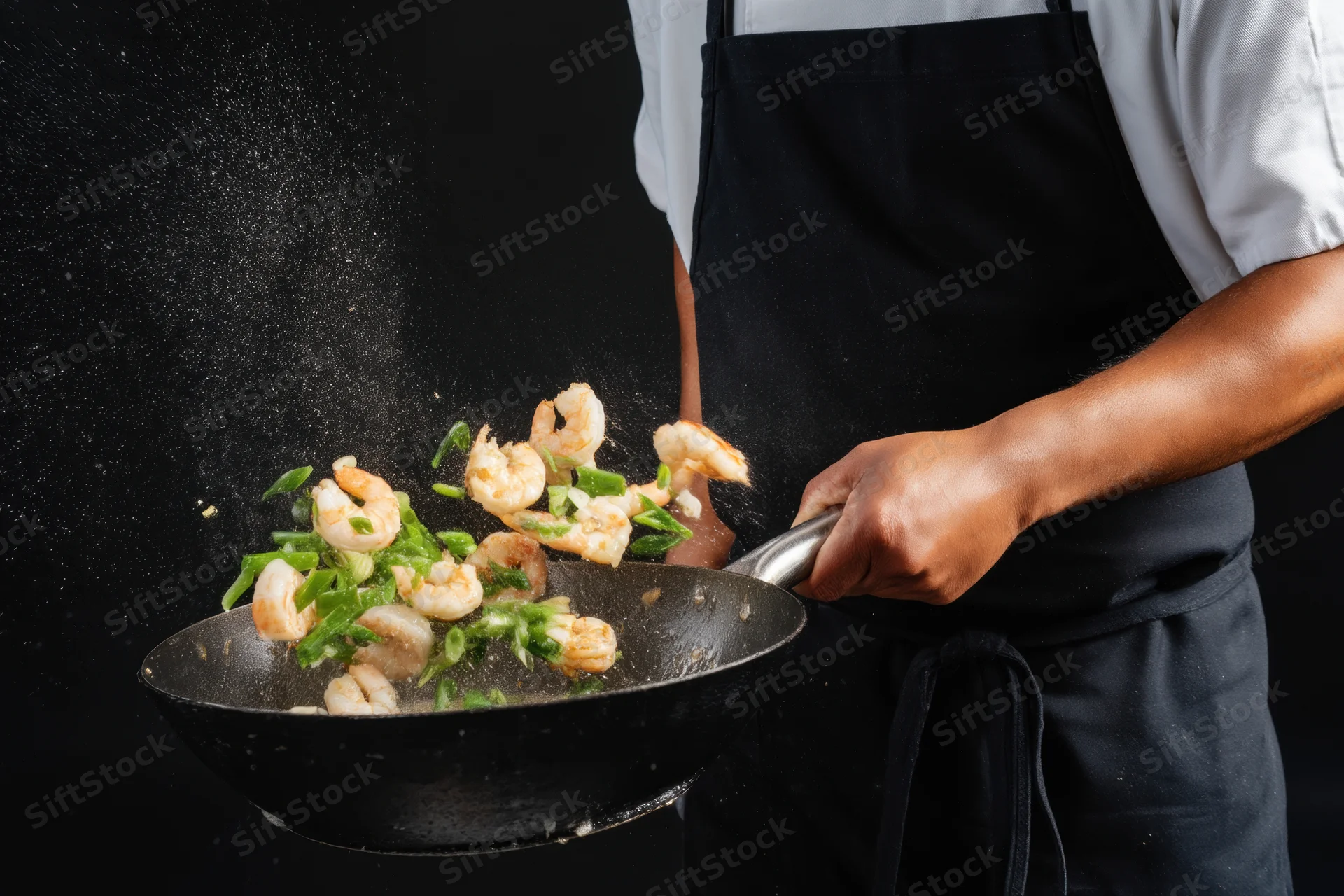Chef tossing shrimp and green onions in a wok against black background
