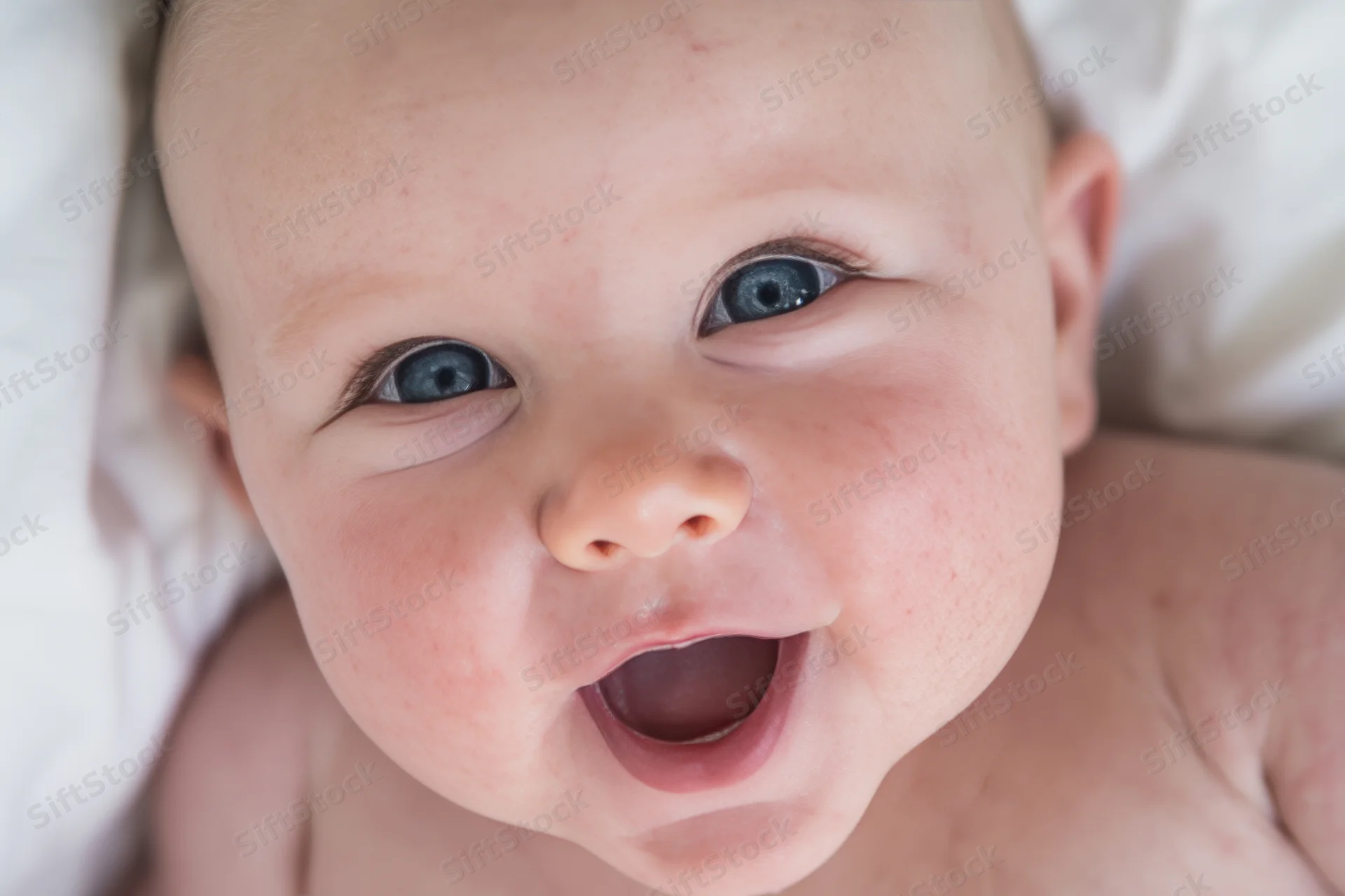 Close-up of a smiling baby with bright blue eyes lying on a white blanket