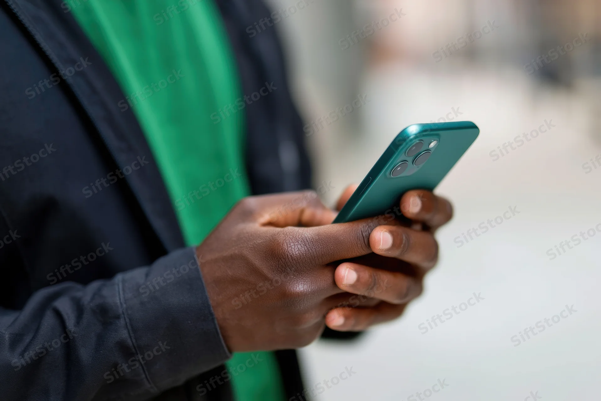 Man holding a smartphone outdoors in a dark jacket and green shirt