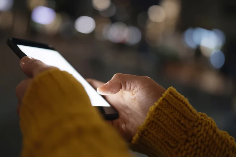 Hands holding smartphone with bright screen against blurred city lights at night