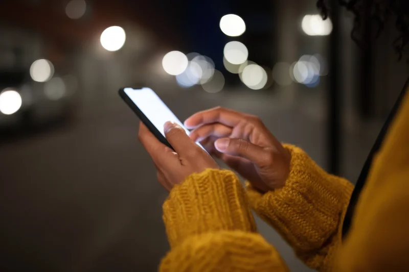 Woman using smartphone at night in city street, hands visible in yellow knit sweater