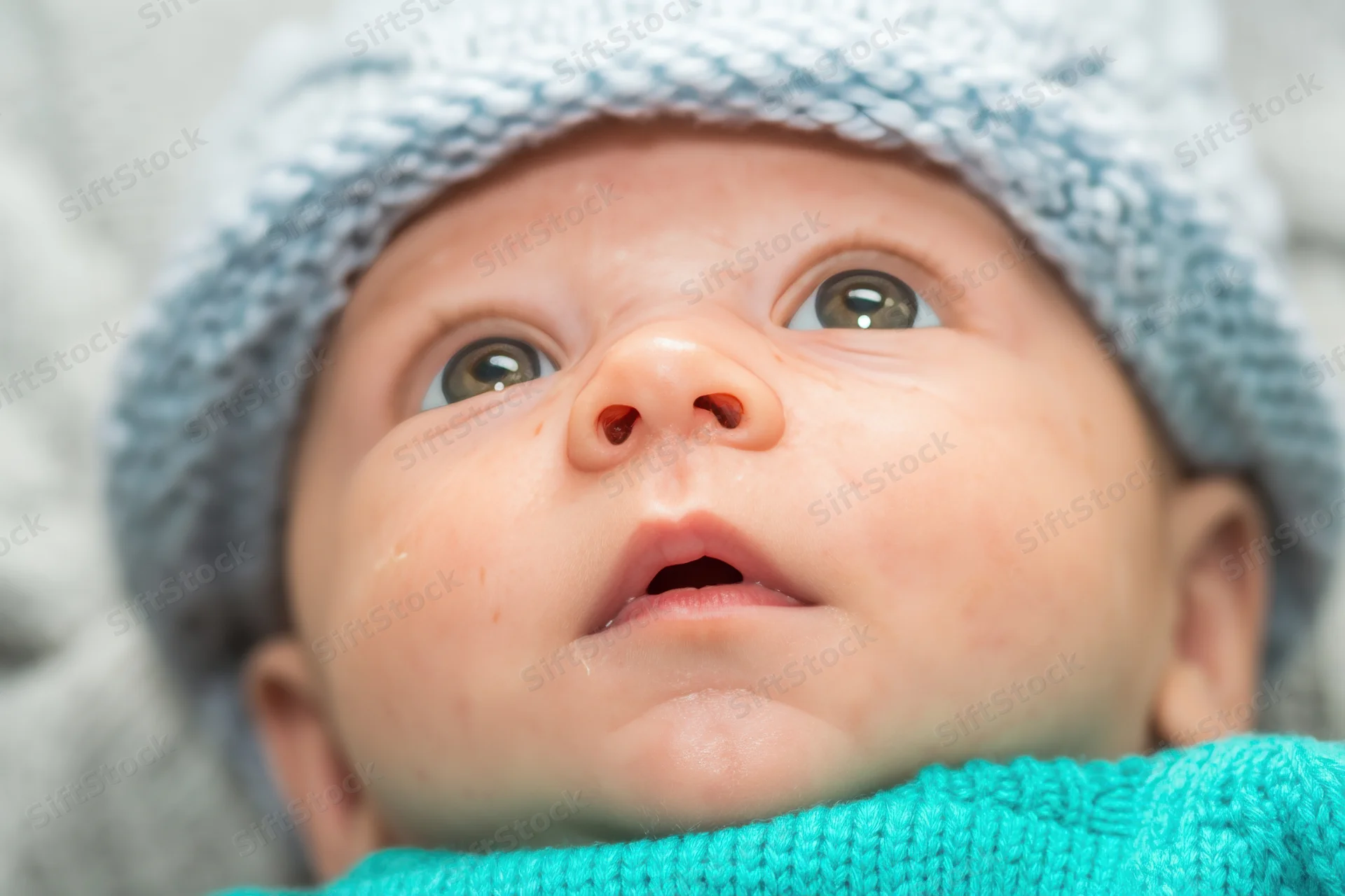 Close-up of a newborn baby wearing a light blue knit hat and turquoise sweater, looking upward with wide eyes