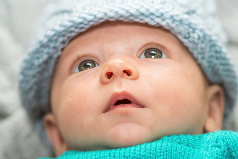 Close-up of a newborn baby wearing a light blue knit hat and turquoise sweater, looking upward with wide eyes