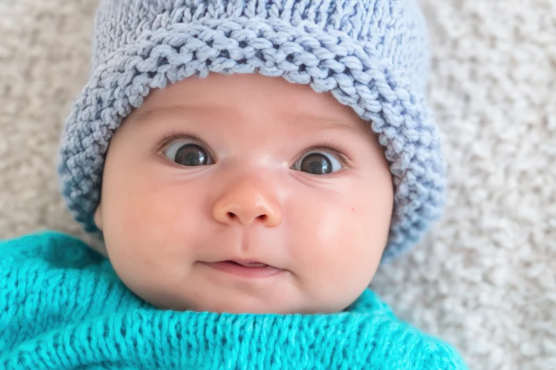 Baby wearing a light blue knit hat and turquoise sweater lying on a textured surface