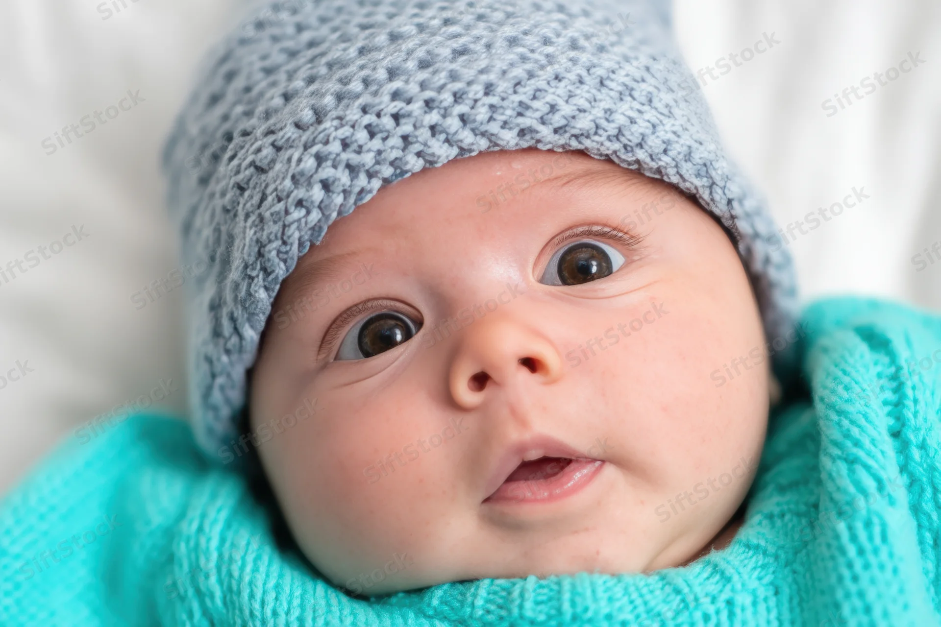 Close-up of a newborn baby wearing a light blue knit hat and turquoise sweater, lying on a white surface