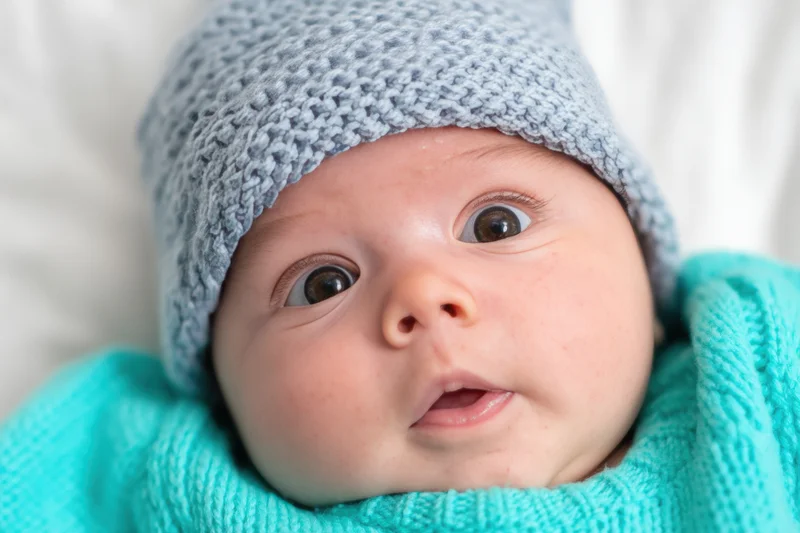 Close-up of a newborn baby wearing a light blue knit hat and turquoise sweater, lying on a white surface