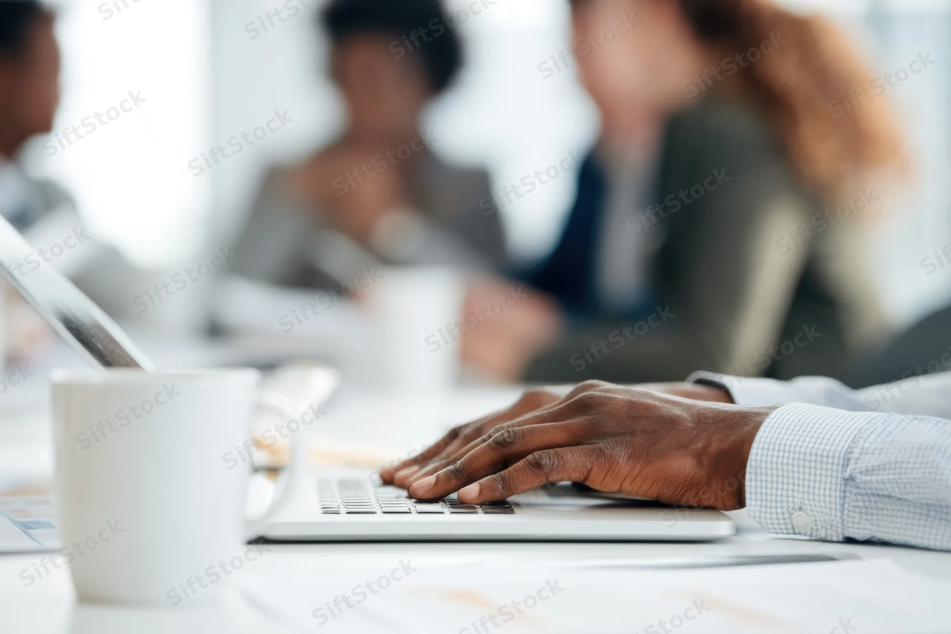 Hands typing on laptop at conference table with colleagues in background