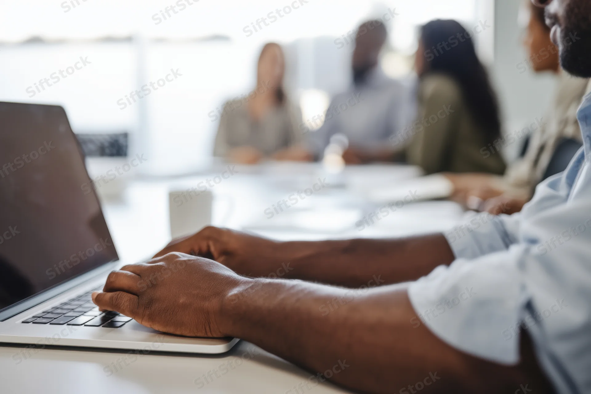 Close-up of hands typing on a laptop at a conference table with colleagues in the background