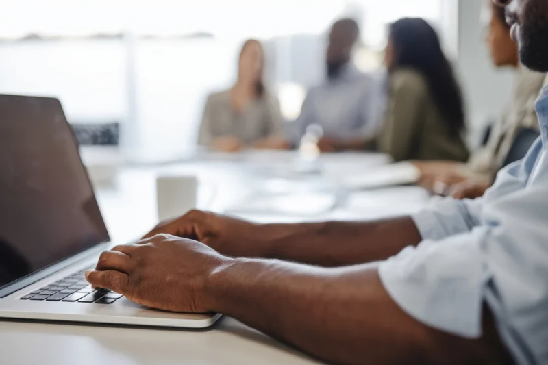 Close-up of hands typing on a laptop at a conference table with colleagues in the background