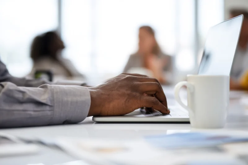 Close-up of hands typing on laptop at conference table with mug and documents