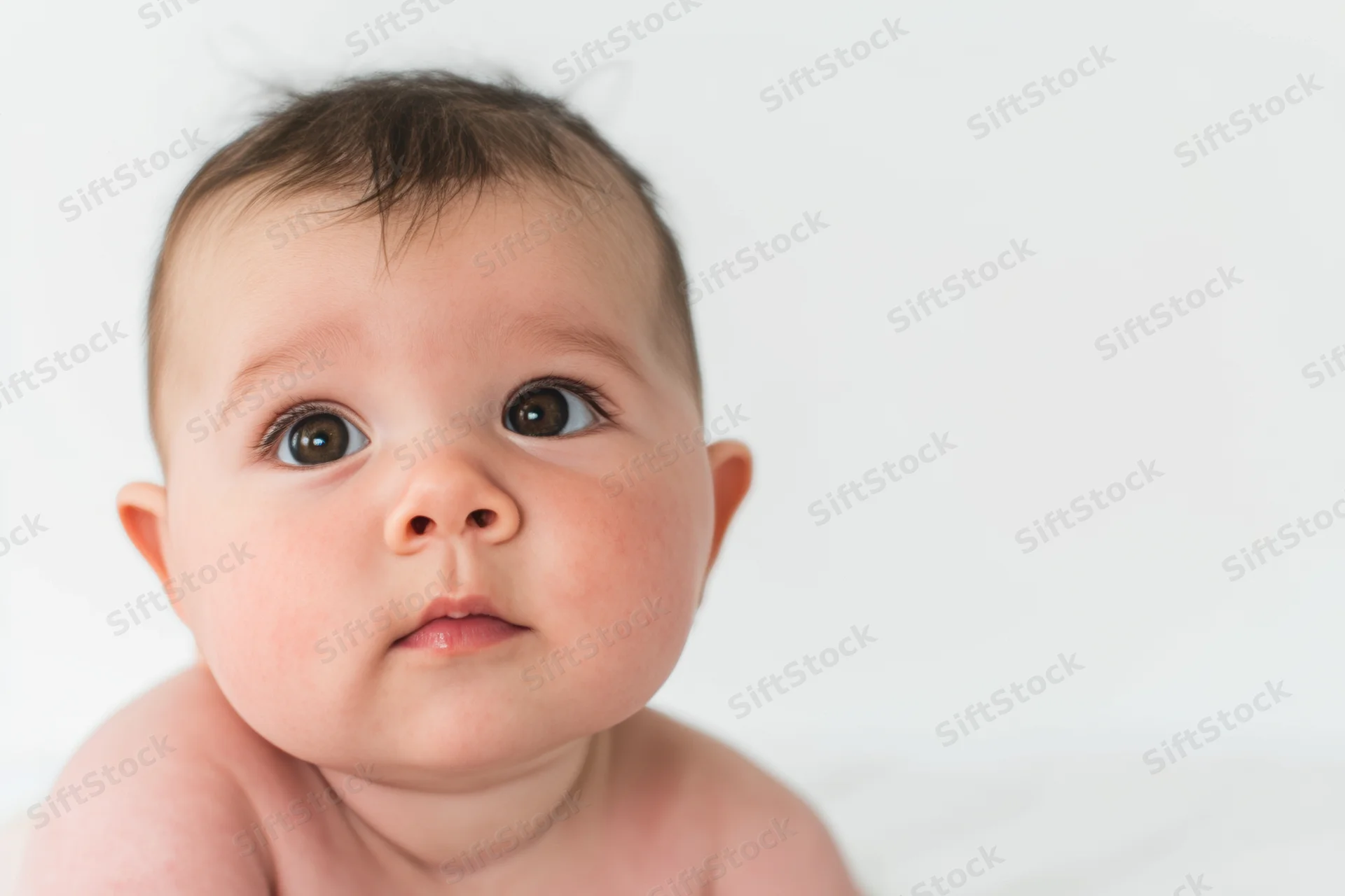 Close-up portrait of a baby with rosy cheeks and soft skin looking up against a white background