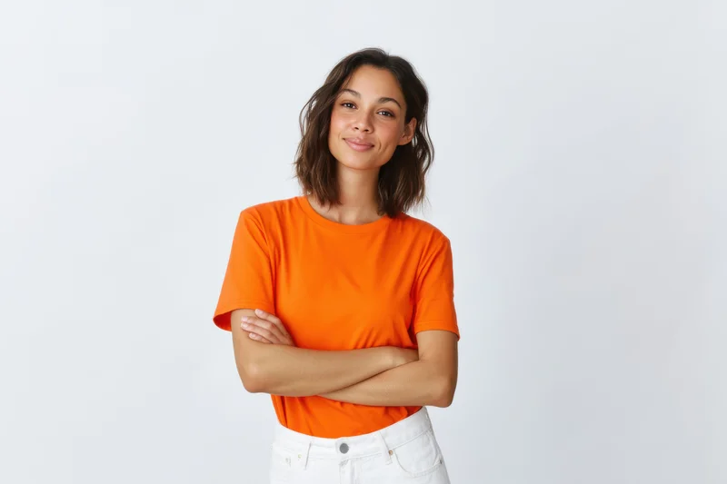 Young woman with arms crossed smiling in orange tshirt and white jeans against white background
