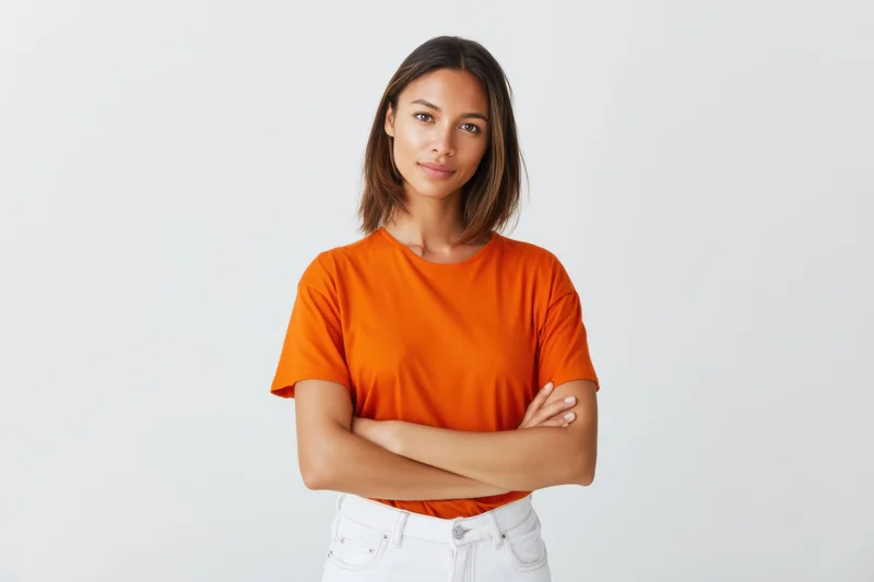 Confident young woman with arms crossed wearing orange tshirt and white jeans in studio setting