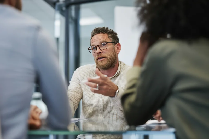 Man with glasses and beard talking and gesturing during a meeting in a modern office with colleagues around a glass table