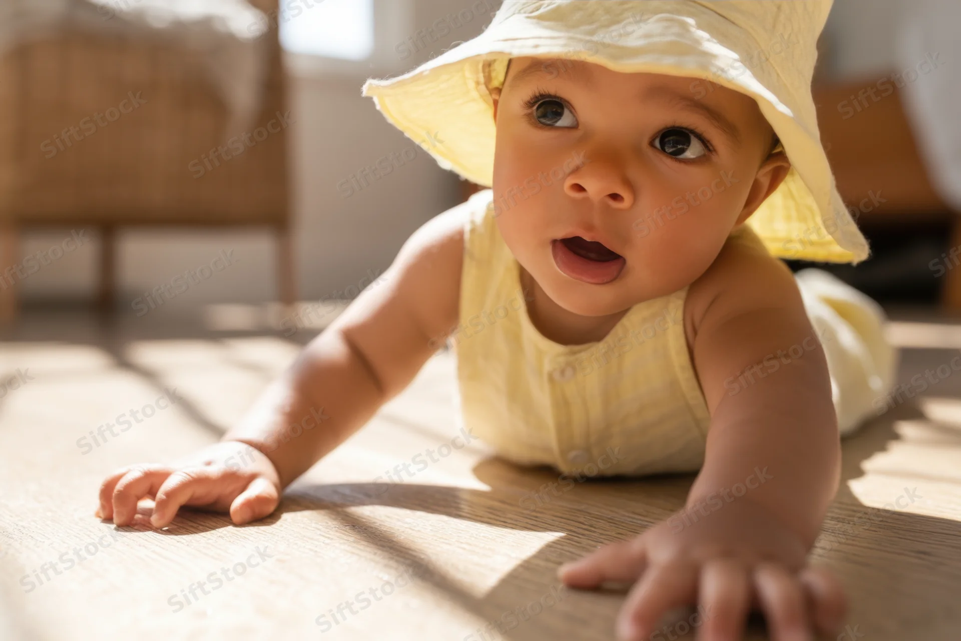 Baby crawling on sunlit wooden floor wearing yellow hat and outfit
