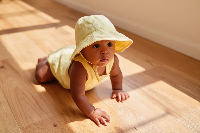 Baby crawling on sunlit wooden floor wearing yellow bucket hat