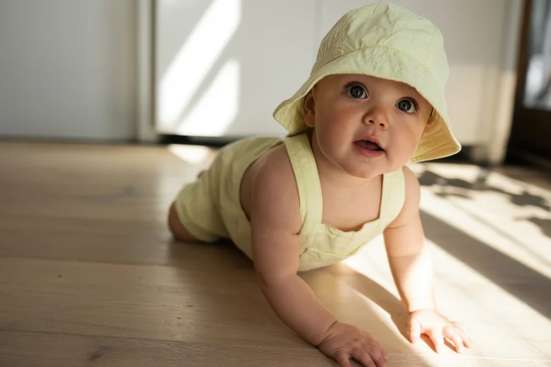 Baby crawling on sunlit wooden floor wearing yellow hat and outfit