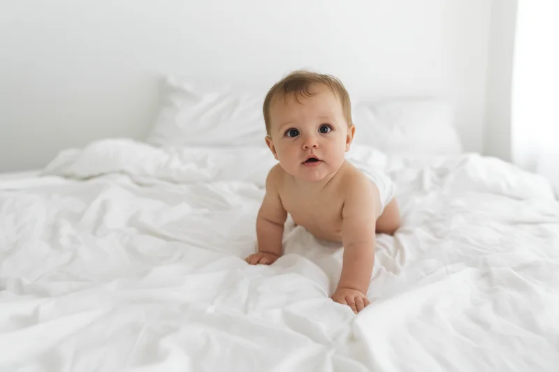 Baby crawling on white bed in diaper, looking at camera, in bedroom