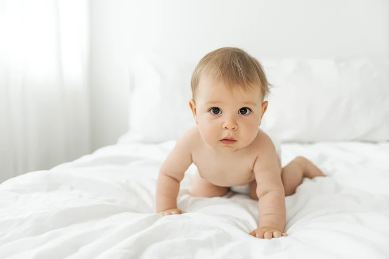 Baby crawling on white bed in bedroom, looking at camera