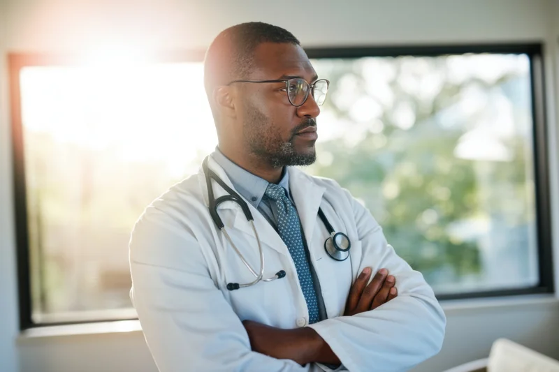 Male doctor in white coat with stethoscope around neck, arms crossed, standing by window in clinic