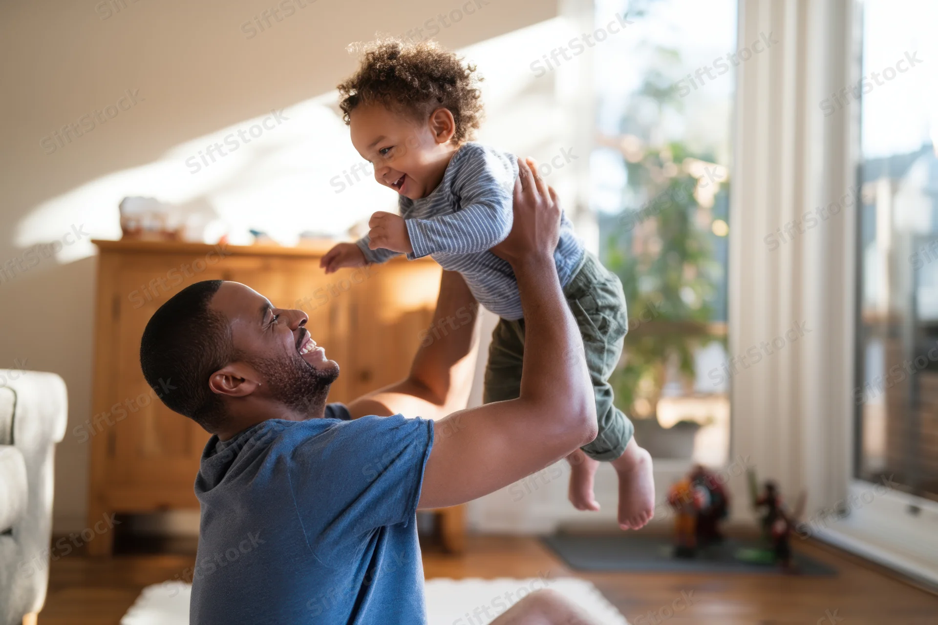 Father lifting young son in living room, both smiling, sunlight streaming through window