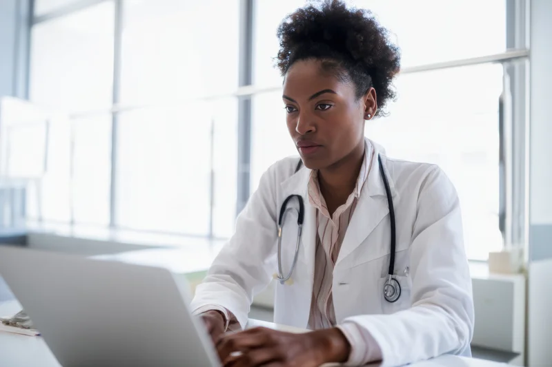 Female doctor in white coat typing on laptop at bright office desk
