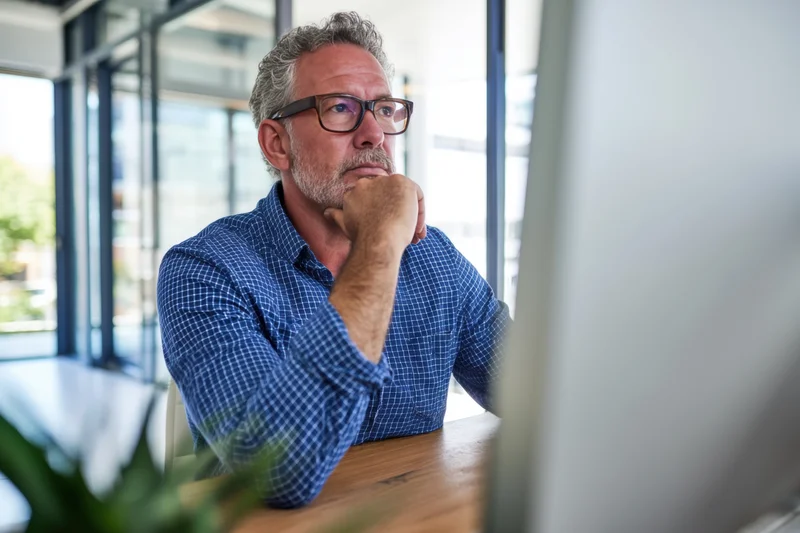 Man in blue checkered shirt and glasses concentrating at computer desk in office