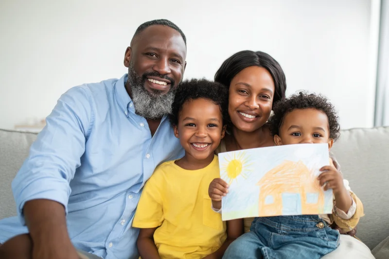 A black family of four smiling together on a gray sofa while a child holds up a crayon drawing of a house and sun