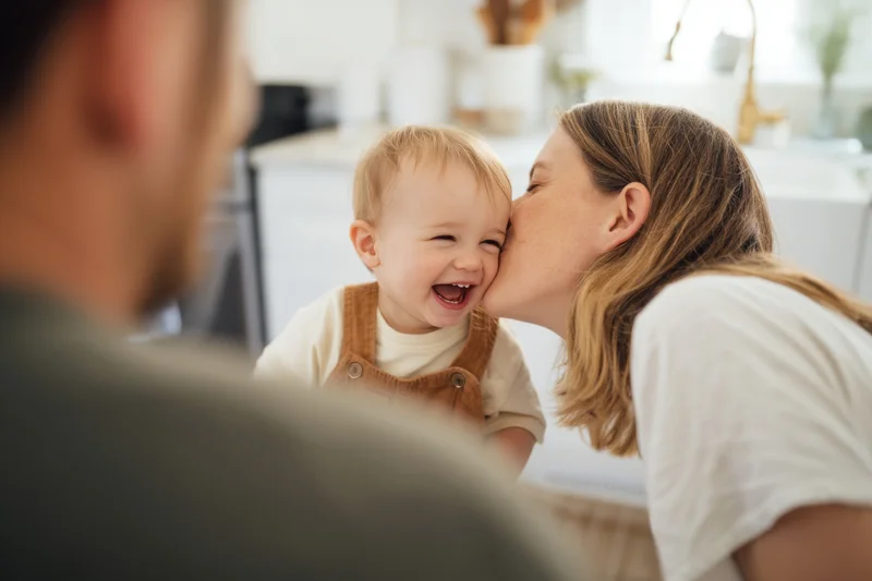 Mother kissing toddler on the cheek in a bright kitchen with father in foreground