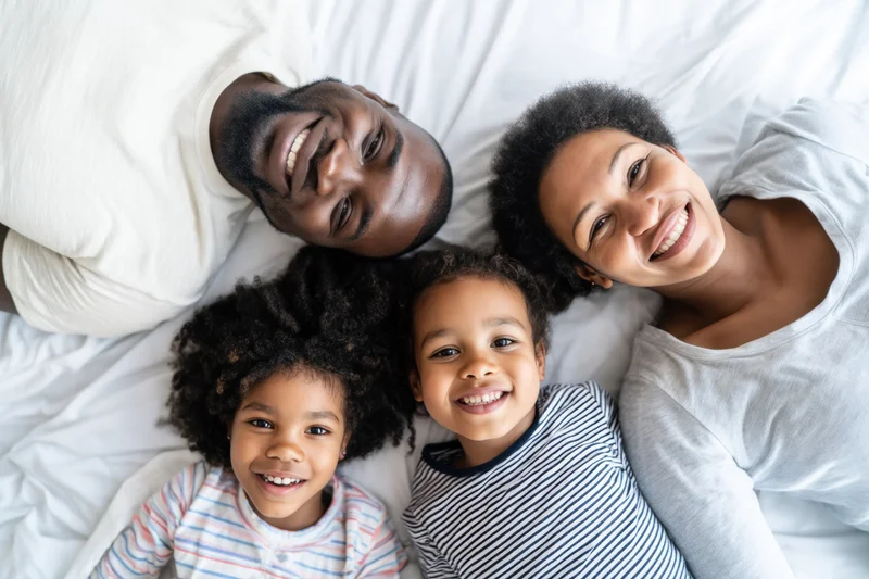Family of four smiling while lying together on a white bed in a bedroom