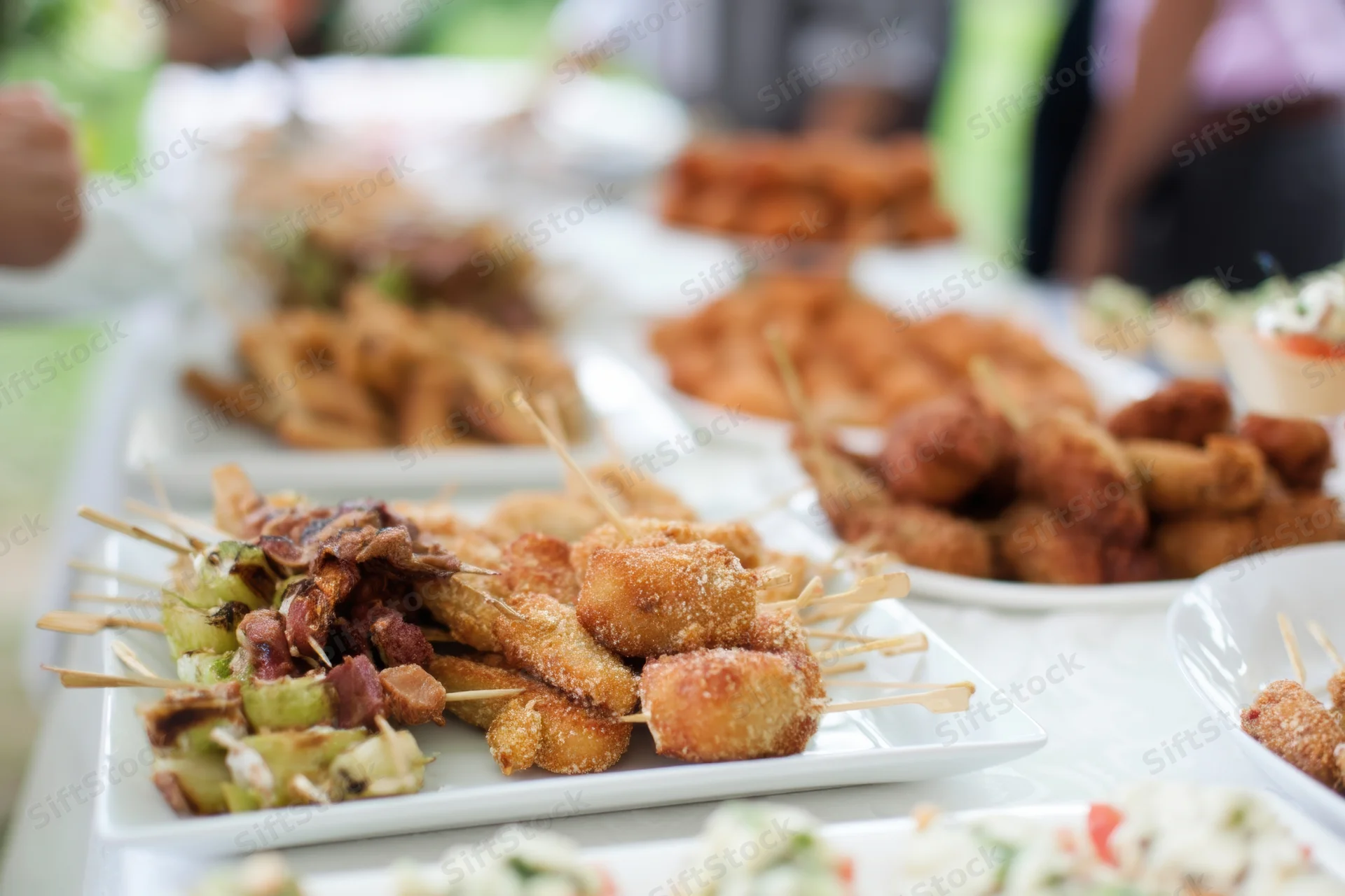 Skewers and croquettes arranged on white plates at a buffet table during a daytime outdoor gathering