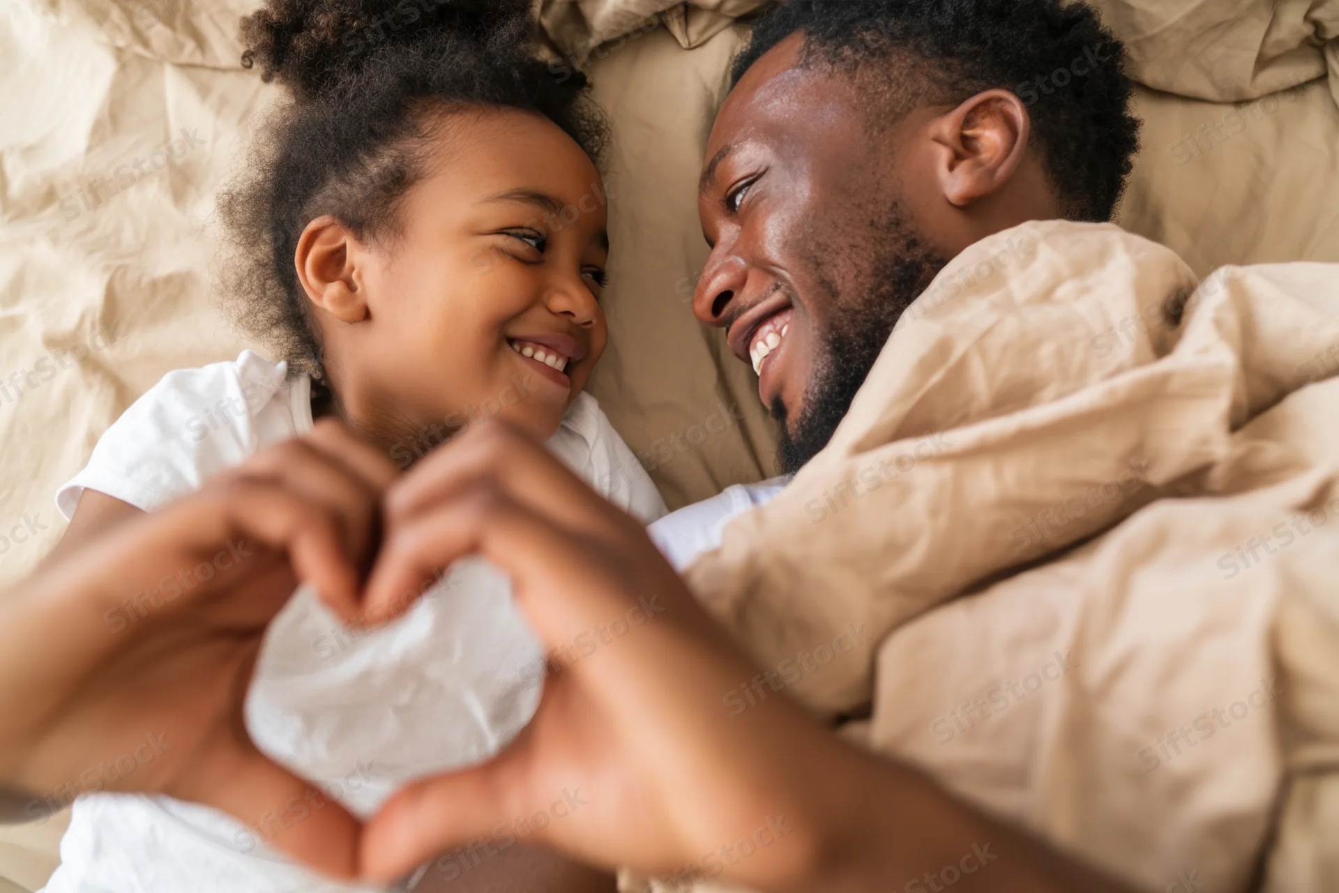 Father and daughter lying in bed smiling at each other while making a heart shape with their hands
