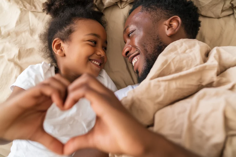 Father and daughter lying in bed smiling at each other while making a heart shape with their hands