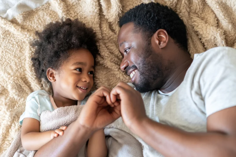 Father and young daughter lying together on a cozy bed, smiling and holding hands, wrapped in a soft blanket