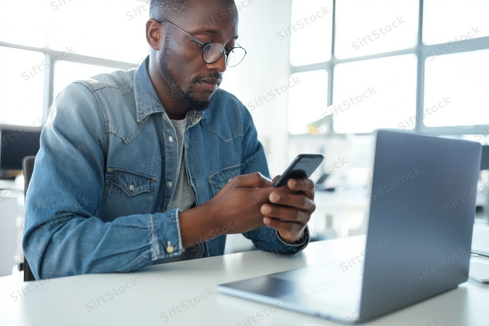 Man using smartphone at desk with laptop in bright office