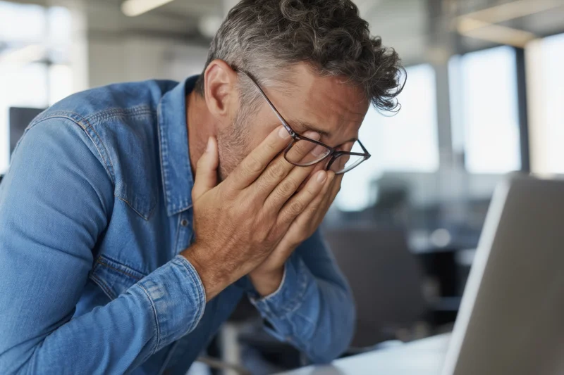 Man in denim shirt covering face with hands at laptop in modern office